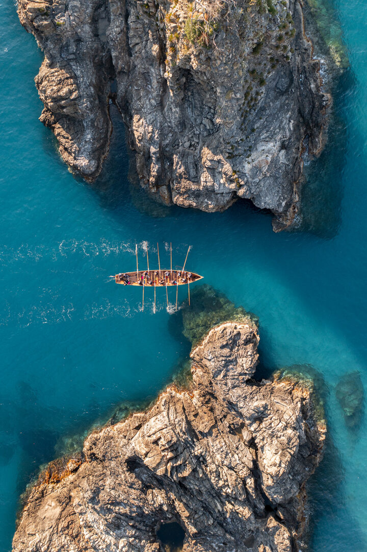 Come Ulisse tenta di superare i mostri di Scilla e Cariddi, la riproduzione di una storica imbarcazione in legno attraversa lo scoglio dell’ulivo al tramonto. Ulivarella di Palmi, Costa Viola.